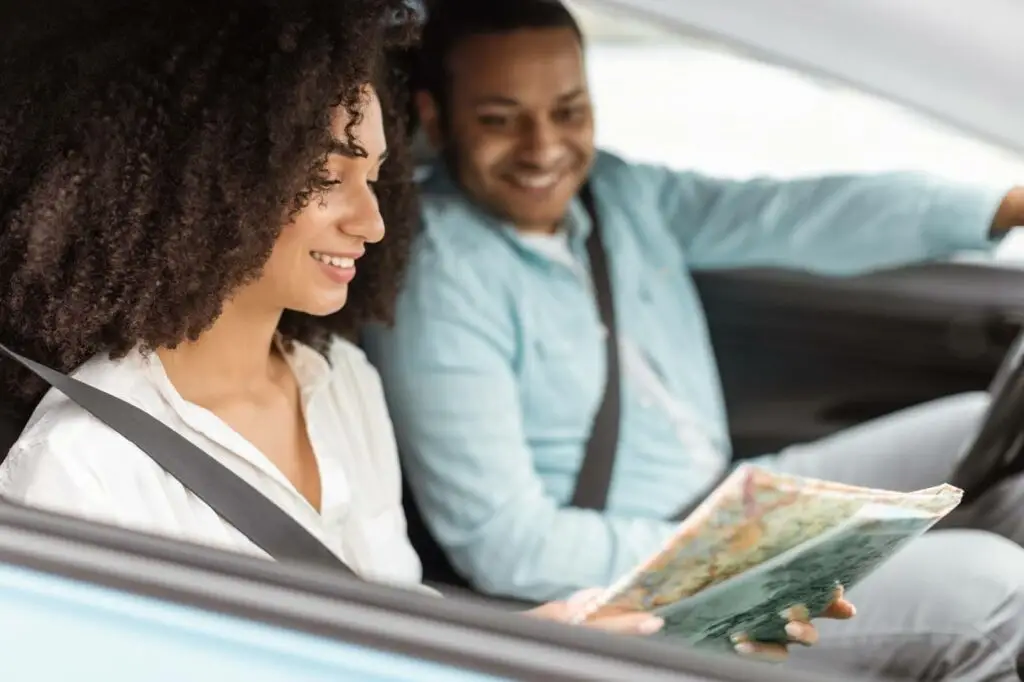Smiling woman in the passenger seat reads a map while a man drives, both wearing seatbelts inside a car—an ideal scene for efficient routes managed with Transportation Dispatch Software.