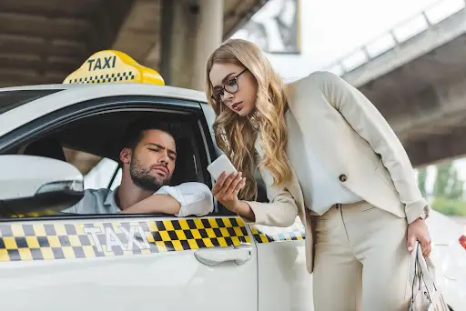 A woman in a beige suit shows her phone, featuring PerGO NEMT Software, to a male taxi driver leaning out of the window under an overpass. The driver looks at the screen with a serious expression.