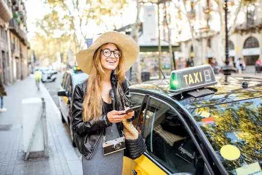 A smiling woman in a sunhat, glasses, and black jacket stands next to a yellow taxi on a city street, using her phone—perhaps checking transportation dispatch software—with trees and buildings in the background.