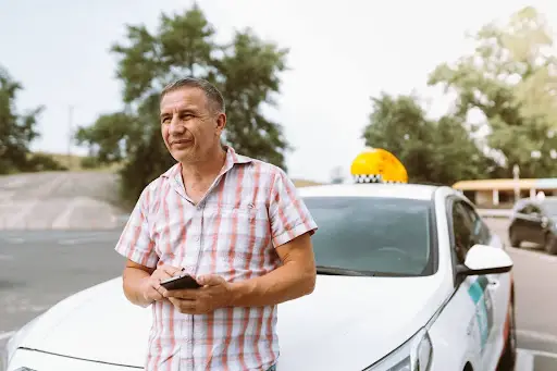 A man in a plaid shirt stands in front of a white taxi, holding a smartphone. Trees and a road are visible in the background on a sunny day, highlighting the convenience of NEMT Software like PerGo for seamless transportation management.