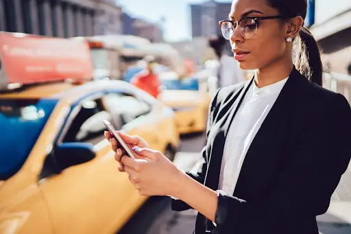 A woman wearing glasses and a black blazer stands on a city street, looking at her smartphone. A yellow taxi is behind her&mdash;she might be using PerGo transportation dispatch software amid the bustling urban scene.