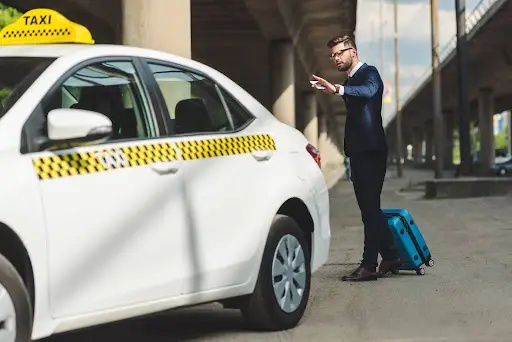 A man in a suit with a blue suitcase stands on a sidewalk, reaching his hand out toward a white taxi&mdash;efficiently managed by transportation dispatch software&mdash;parked nearby under an overpass.