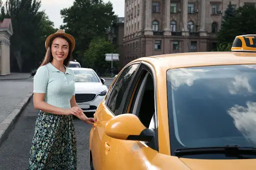 A smiling woman wearing a hat and floral skirt stands on a city street, about to enter a yellow taxi cab managed with advanced Transportation management Software. Trees and buildings are visible in the background.