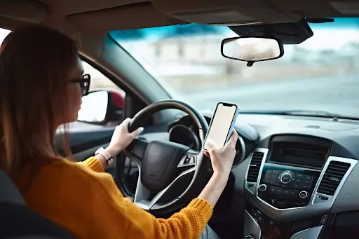 A woman wearing glasses and an orange sweater is driving a car while using PerGo, a leading transportation dispatch software, on her smartphone. The road is visible through the windshield.