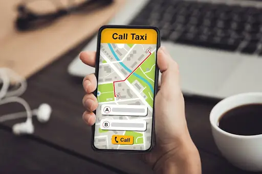 A hand holds a smartphone with the PerGo taxi app open, displaying a map, route, and &ldquo;Call Taxi&rdquo; button. A cup of coffee, a pen, and a laptop&mdash;perhaps running NEMT Software&mdash;sit on the desk in the background.