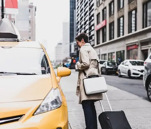 A person wearing a beige trench coat stands on a city street with a suitcase and briefcase, about to get into a yellow taxi. Tall buildings and parked cars line the street.