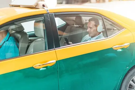 A man wearing headphones sits in the back seat of a yellow and green taxi, looking at his smartphone. The driver is visible in the front seat, wearing a blue shirt.