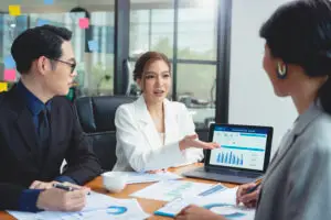 Three business professionals have a meeting at a desk with charts and papers. A woman in a white blazer gestures toward a laptop showing marketing graphs and data, while the others listen and take notes.