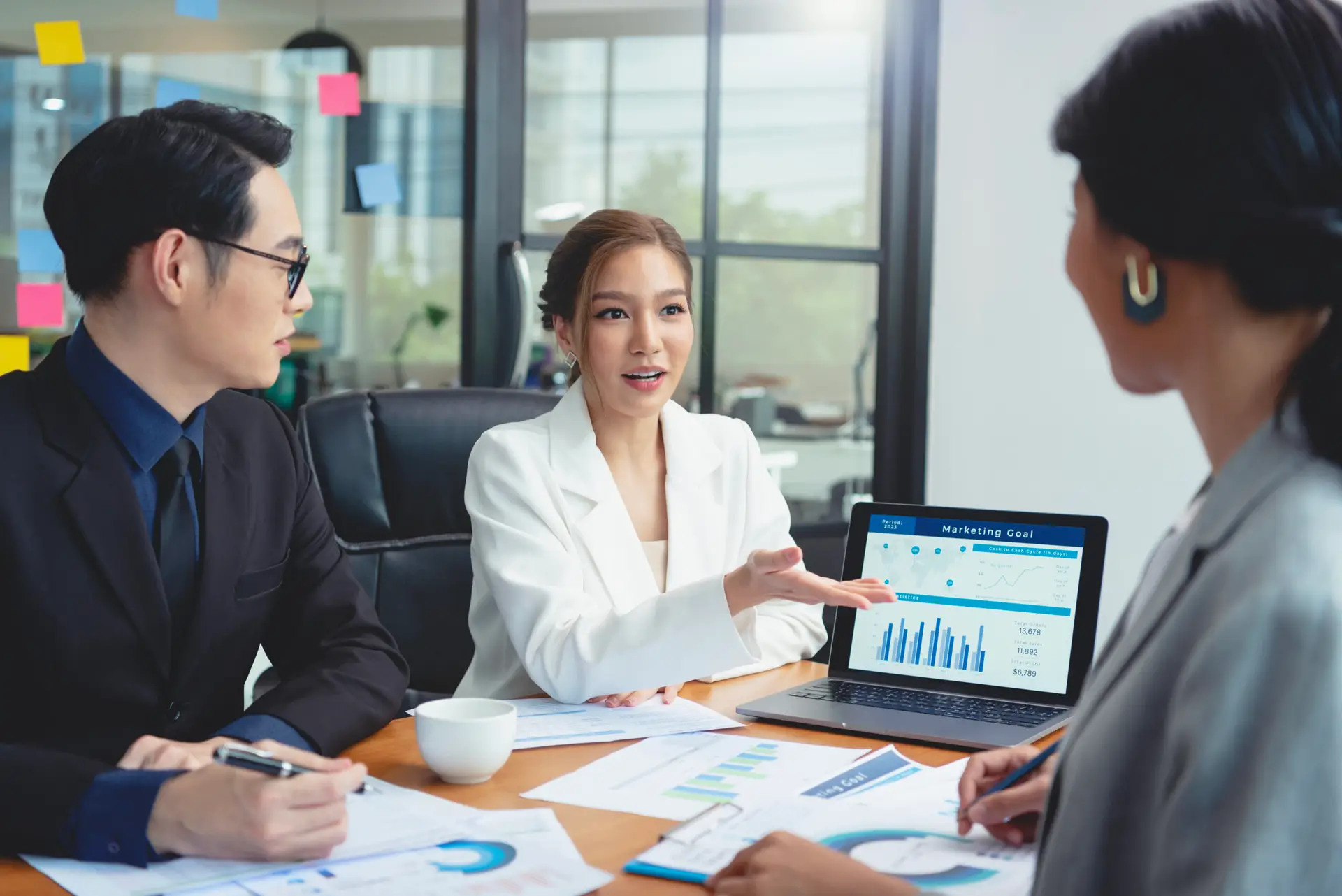 Three business professionals have a meeting at a desk with charts and papers. A woman in a white blazer gestures toward a laptop showing marketing graphs and data, while the others listen and take notes.