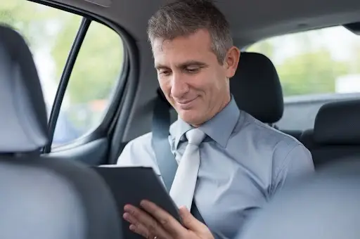 A man in a dress shirt and tie sits in the backseat of a car, wearing a seatbelt and smiling as he looks at a tablet device.