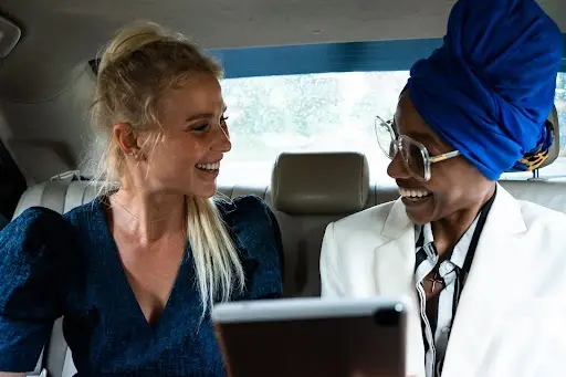 Two people sitting in the backseat of a car, smiling and facing each other. One is wearing a blue dress, the other a white jacket, glasses, and a bright blue headwrap. A tablet is visible in the foreground.
