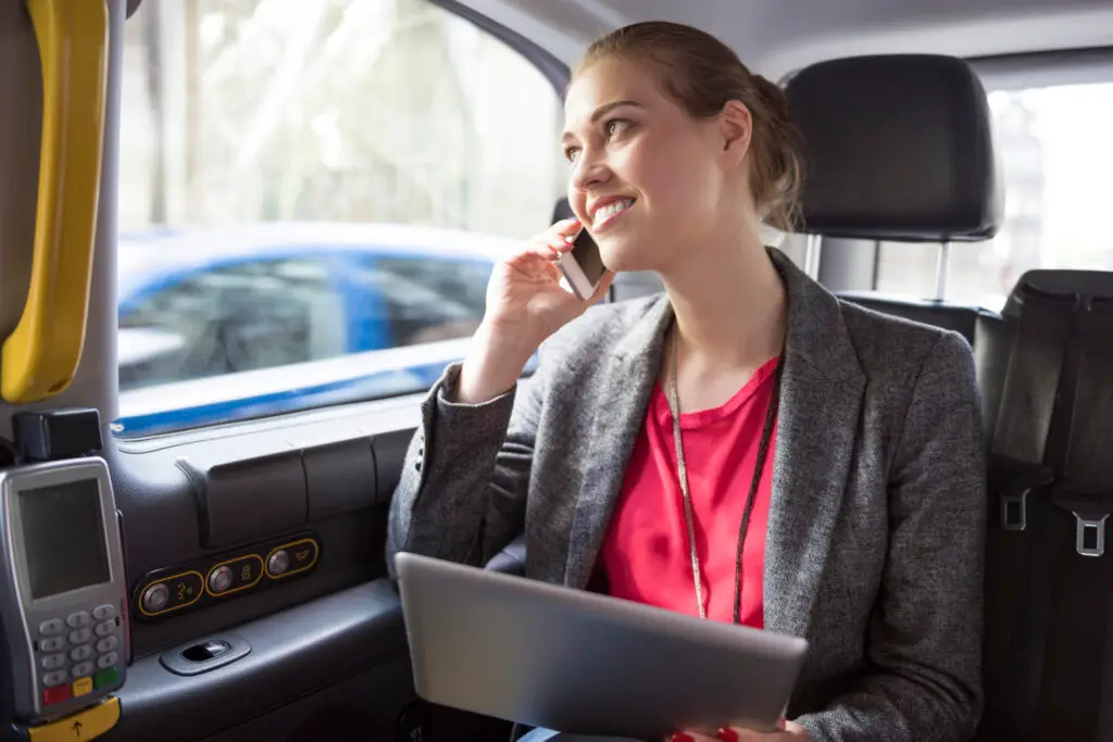 A woman in business attire sits in the backseat of a car, holding a tablet on her lap and smiling while talking on her phone. She appears to be working during her commute.
