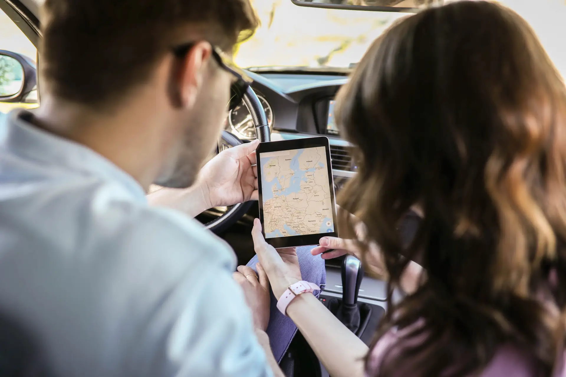 Two people sitting in a car look at a digital map on a tablet, planning a route. The steering wheel and dashboard are visible, and the map on the screen shows part of Europe.