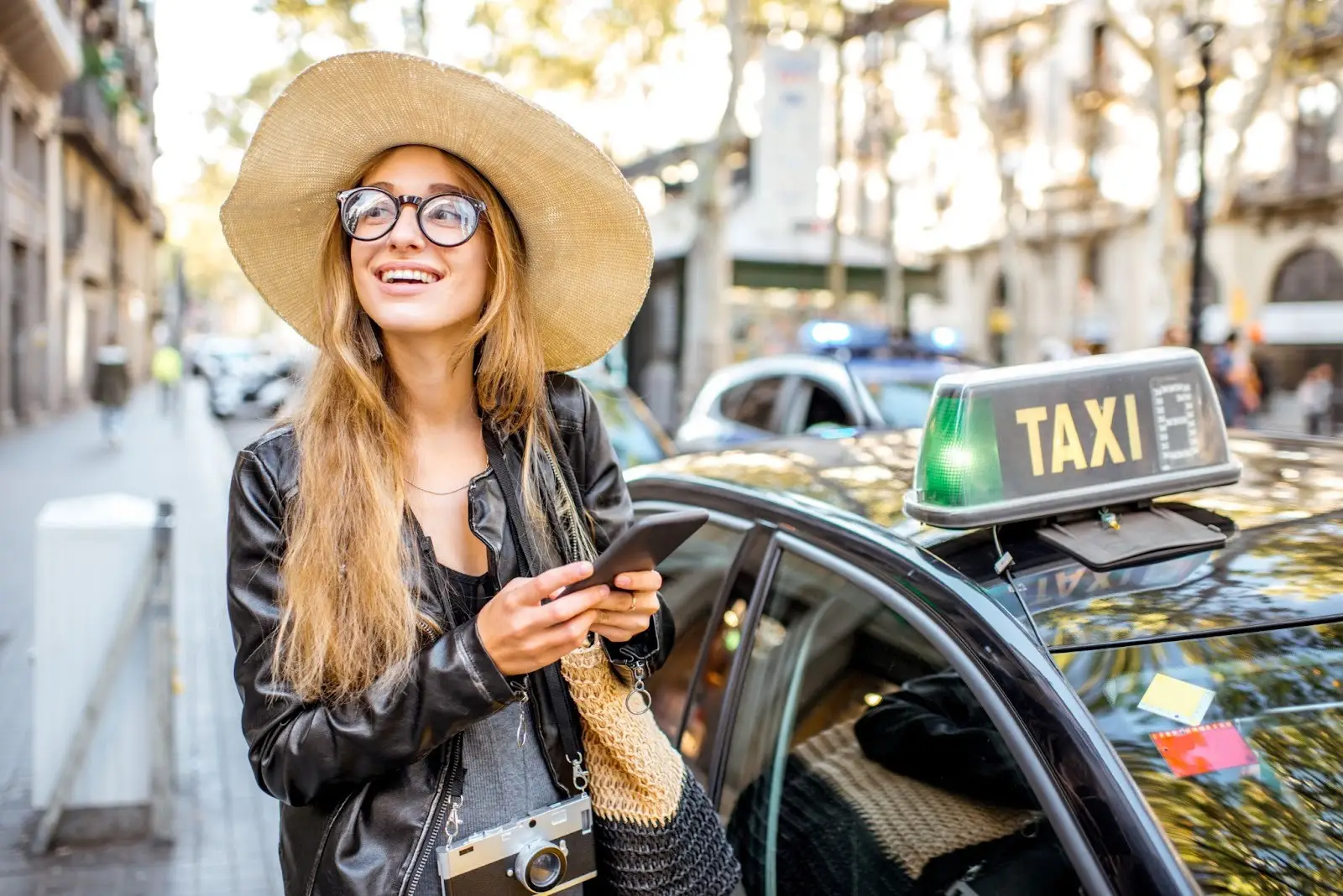 A smiling woman in a large hat and glasses stands by a taxi, holding a smartphone. She wears a leather jacket and camera, standing outdoors on a sunny city street with blurred buildings and cars in the background.