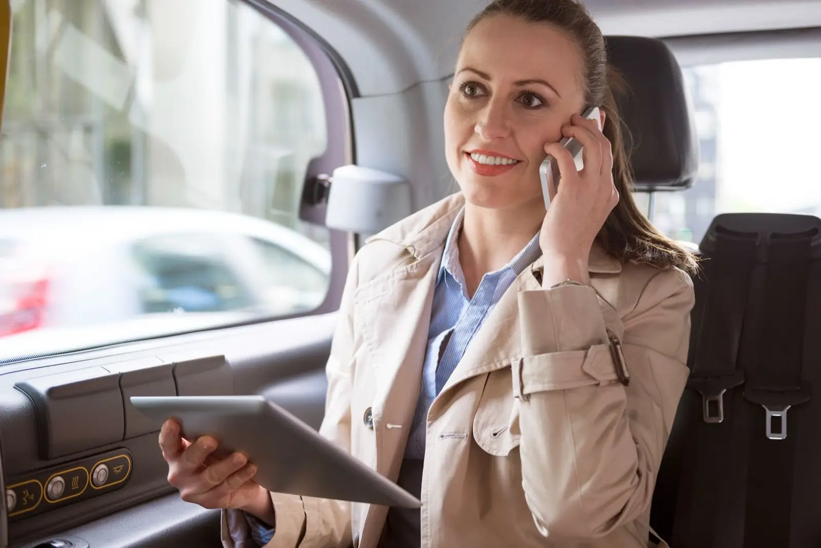 A woman in a beige trench coat sits in the backseat of a car, smiling while talking on a smartphone and holding a tablet in her other hand.