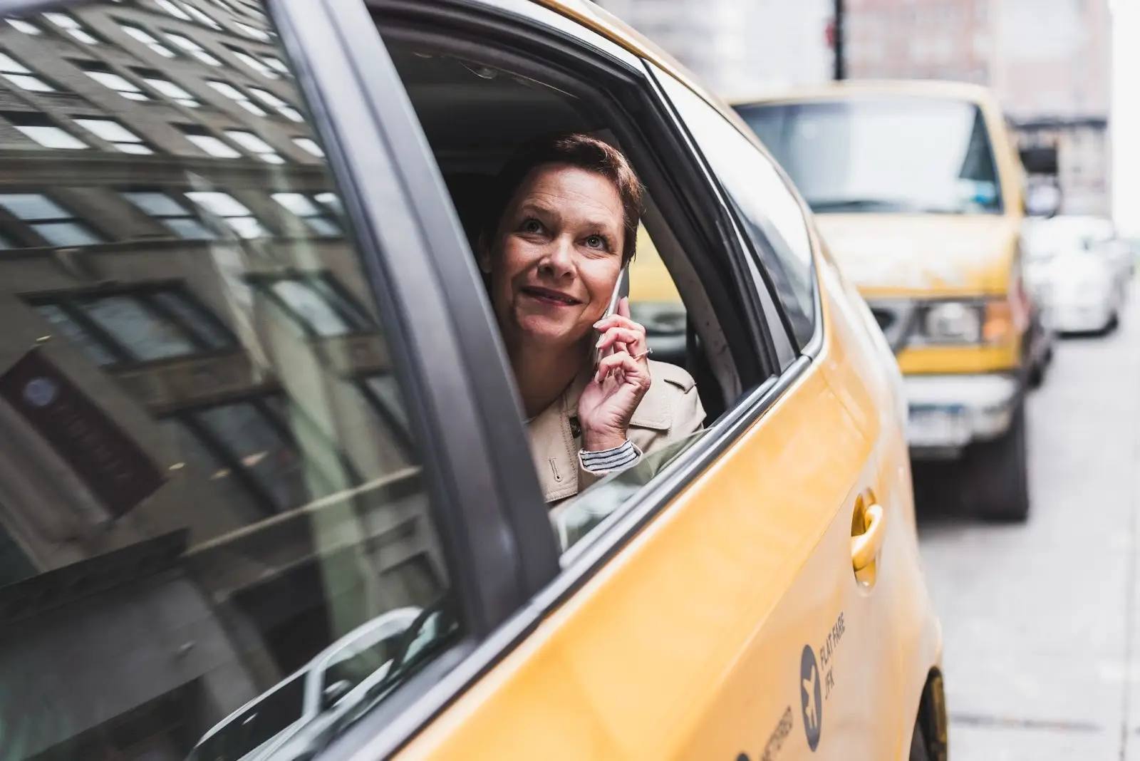 A woman sits in the back seat of a yellow taxi, smiling and talking on her cellphone while looking out the window at city buildings.