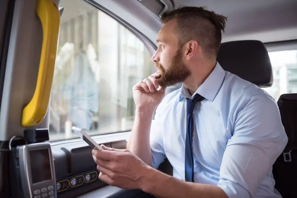 A man in a dress shirt and tie sits in the back seat of a car, holding a smartphone and looking thoughtfully out the window.