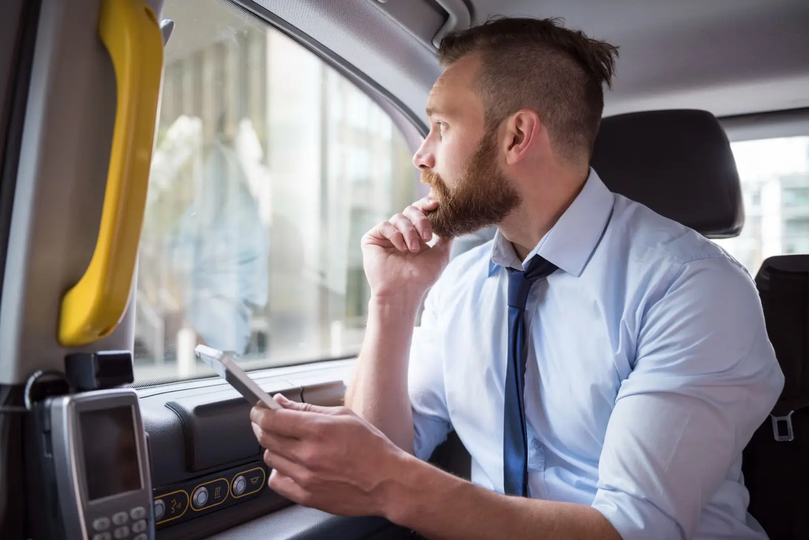 A man in a dress shirt and tie sits in the back seat of a car, holding a smartphone and looking thoughtfully out the window.