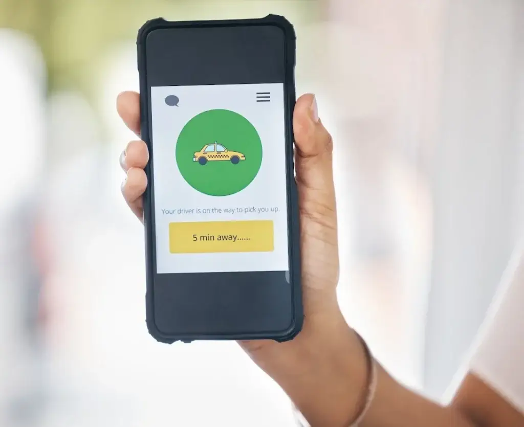 A hand holding a smartphone displaying a taxi app screen with a graphic of a yellow cab and text stating, “Your driver is on the way to pick you up. 5 min away.”.