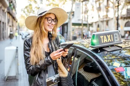 A smiling woman in a hat and glasses stands next to a black taxi on a city street, holding her phone and a woven bag, with a camera hanging from her neck.