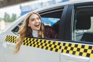 A smiling woman with long blonde hair leans out of the open window of a white taxi with a yellow and black checker pattern on the side.