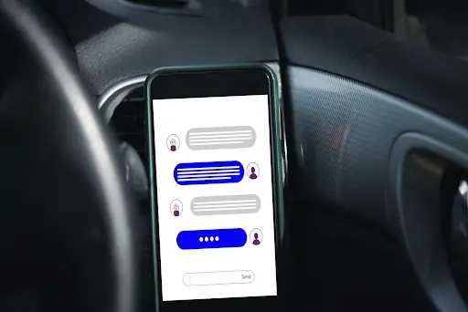 A smartphone mounted in a car displays a chat conversation with blue and gray message bubbles, suggesting someone is texting while driving.