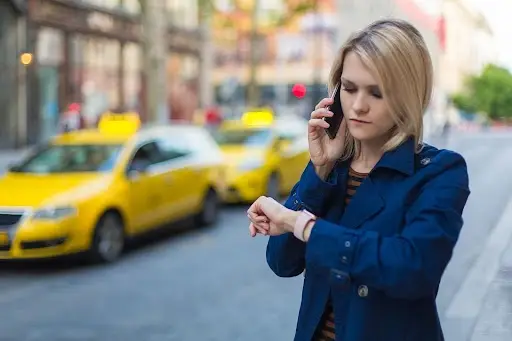 A woman in a blue coat stands on a city street, talking on her phone and looking at her watch. Yellow taxis are visible in the background.