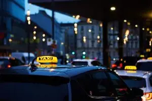 Taxis with illuminated signs are lined up on a city street at dusk, with blurred buildings and streetlights in the background. The atmosphere appears busy and urban.