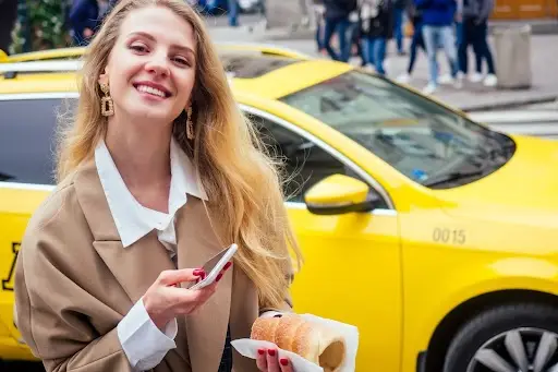 A smiling woman in a beige coat holds a phone and a pastry while standing in front of a yellow taxi on a busy city street.