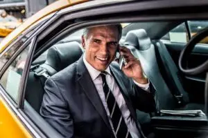 A man in a suit sits in the back seat of a car, smiling while talking on a smartphone. A newspaper is beside him, and the car door is open, showing part of a yellow vehicle exterior.