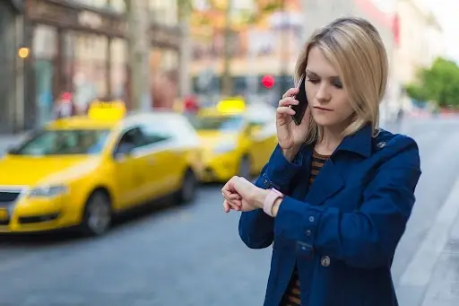 A woman with blonde hair stands on a city street, talking on her phone and checking her watch. Two yellow taxis are parked in the background. She appears to be waiting or in a hurry.