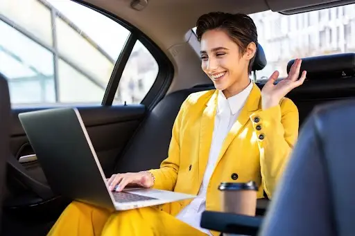 A person in a bright yellow suit sits in the backseat of a car, smiling and waving at their laptop screen during a video call. A coffee cup is next to them. Sunlight streams through the windows.