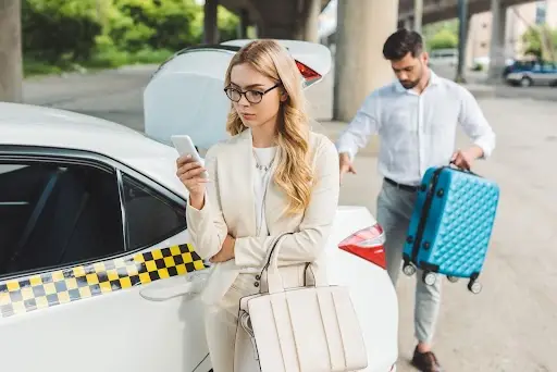 A woman in business attire checks her phone while standing by a white taxi with an open trunk. A man in the background retrieves a blue suitcase from the trunk.