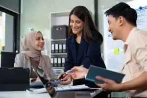 Three colleagues, including a woman in a hijab, a woman in a blazer, and a man, collaborate cheerfully at a desk with laptops, documents, and notebooks in a modern office setting.