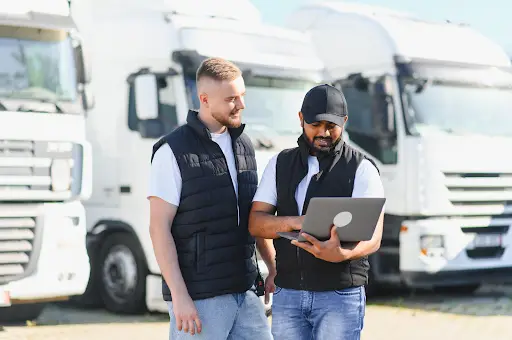 Two men in casual vests stand in front of white semi-trucks. One man holds a laptop, and they both appear to be discussing something on the screen, possibly related to logistics or fleet management.