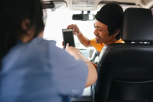 A person in the passenger seat reaches for a smartphone held by a smiling person in the drivers seat inside a car. The focus is on their interaction with the phone.