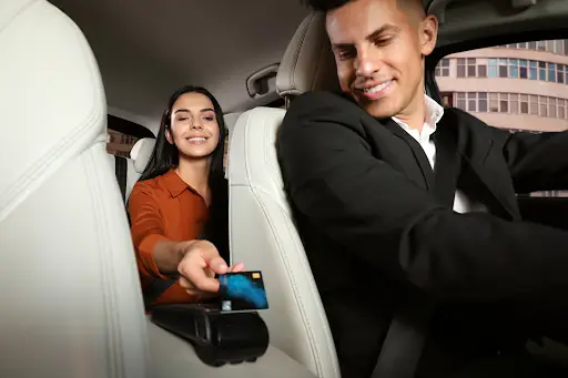 A woman in the back seat of a car hands a credit card to a smiling male driver, who is wearing a suit and sitting in the front seat. They appear to be completing a payment for a ride.