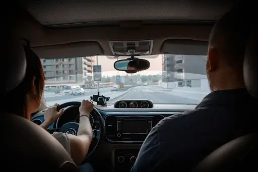View from the backseat of a car showing two people in the front seats, one driving and another in the passenger seat, traveling through an urban area with buildings visible outside.