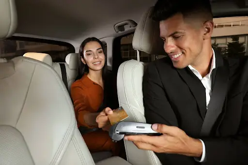 A woman sitting in the back seat of a car hands a credit card to a smiling man in the front passenger seat, who holds a payment terminal. Both appear to be engaged in a ride service transaction.
