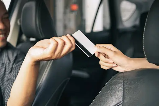 A person in the driver&rsquo;s seat of a car hands a credit card to another person seated next to them, with both partially visible and the car interior in the background.