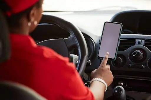 A person wearing a red shirt and cap sits in a car, using their finger to touch a smartphone mounted on the dashboard. The cars steering wheel, controls, and air vents are visible.