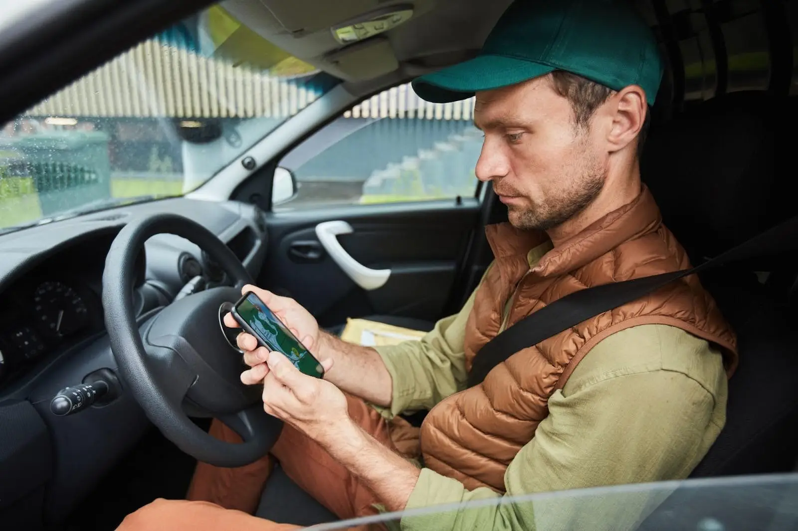 A man wearing a green cap and brown vest sits in the driver&rsquo;s seat of a car, looking at and using his smartphone. The car is parked, and he appears focused on the phone screen.