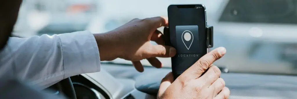 A person in a white shirt adjusts a smartphone in a car mount on the dashboard. The phone screen displays a location pin icon, indicating a navigation or GPS app is open.