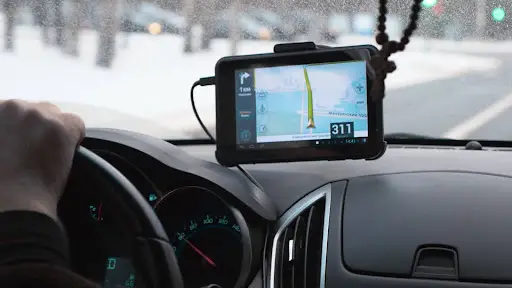 A driver&rsquo;s hand is on the steering wheel while a GPS navigation device on the dashboard displays directions, showing a speed of 31 km/h. Snow is visible outside through the windshield.