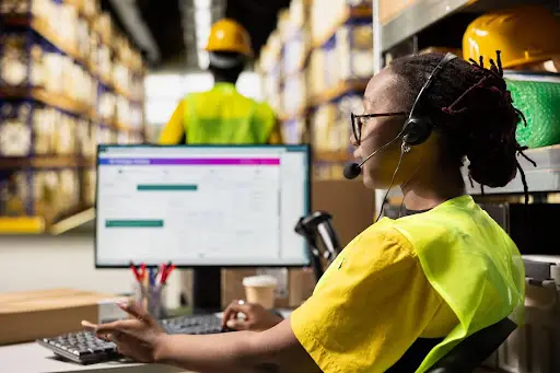A person wearing a headset and yellow safety vest works at a computer in a warehouse, with shelves of boxes in the background and another worker in safety gear visible further down the aisle.